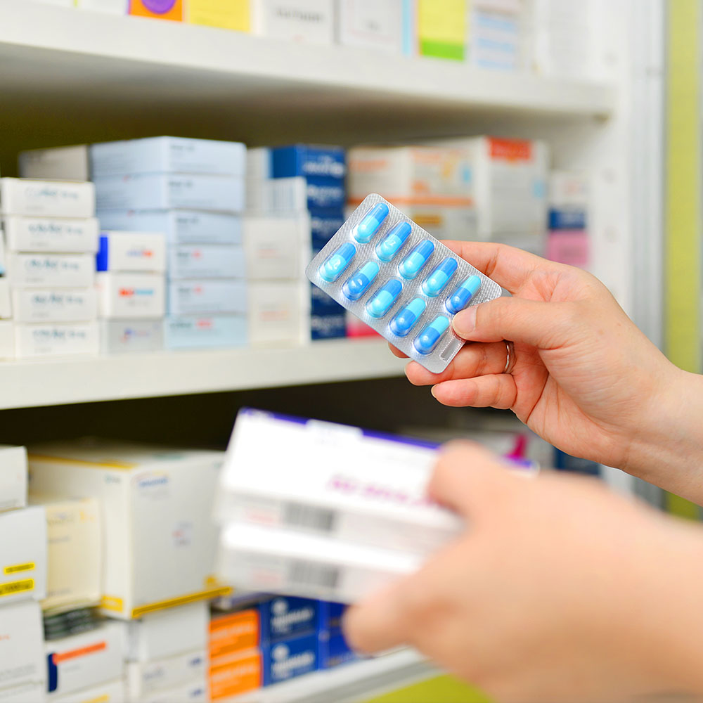 Hand holding packet of blue medication with pharmacy shelves in background